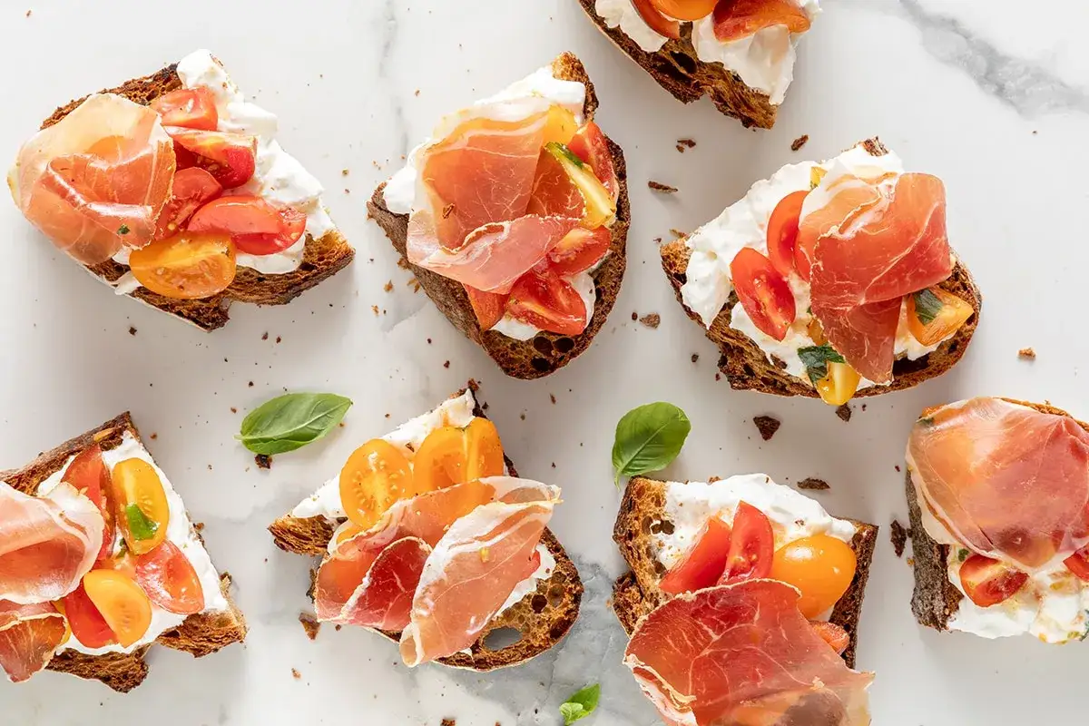 Deliziose tartine per aperitivo con pane tostato, formaggio cremoso, pomodorini colorati e fette di prosciutto crudo.