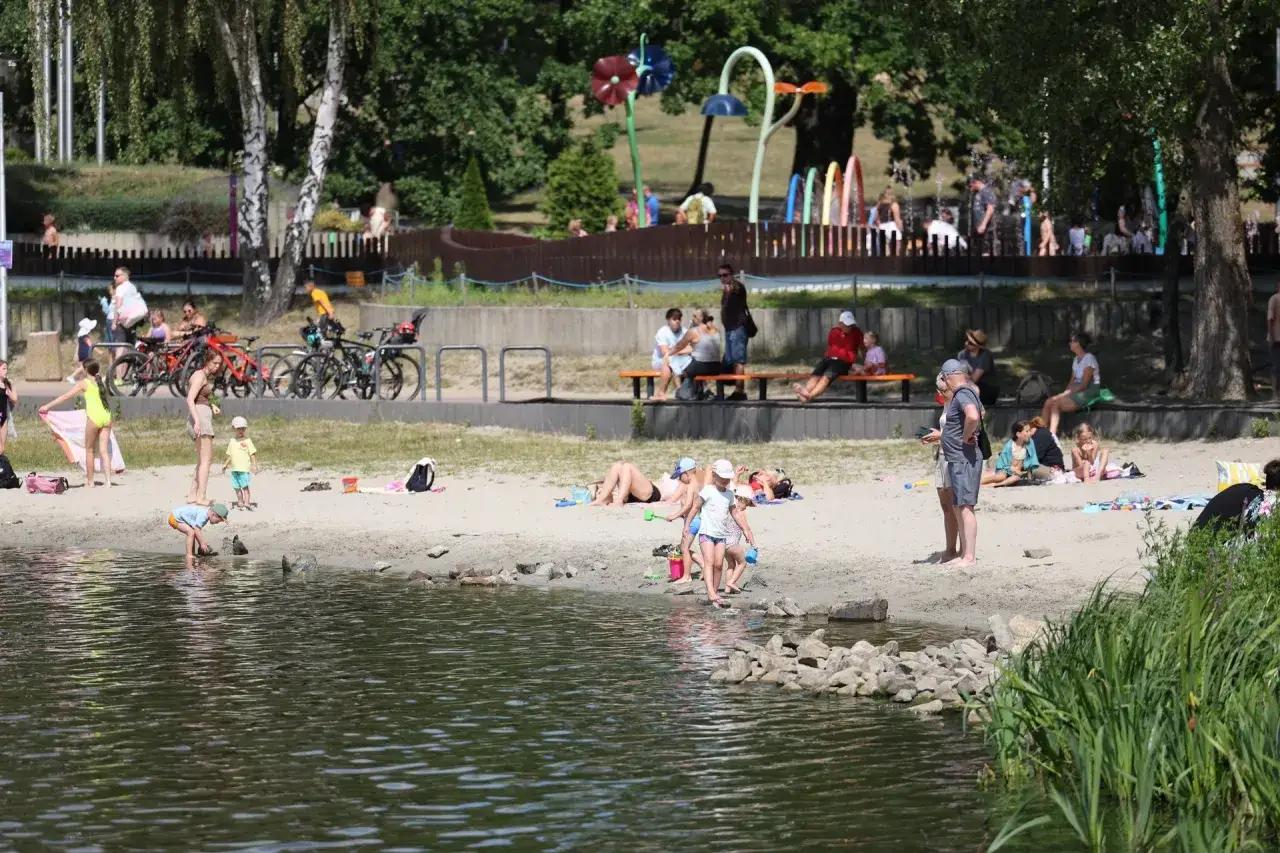 Ludzie odpoczywają na plaży nad jeziorem Paprocany. Dzieci bawią się w wodzie, dorośli opalają się i siedzą na ławkach.