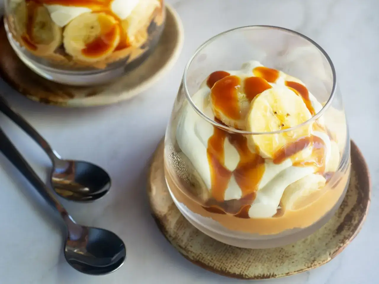 Pyszny deser banoffee w szklance, z bananami, bitą śmietaną i karmelem. Dwa pucharki i łyżeczki na stole.