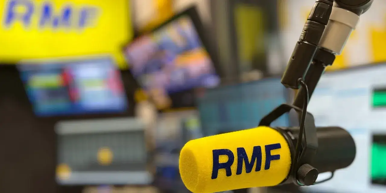 Mikrofon z logo RMF FM w studiu radiowym. Słychać głosy i muzykę, a na ekranach w tle widać wykresy.