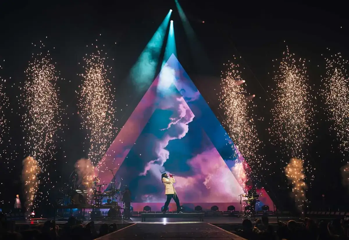 Koncert Thirty Seconds to Mars. Wokalista na scenie, fajerwerki, trójkątny ekran z chmurami.