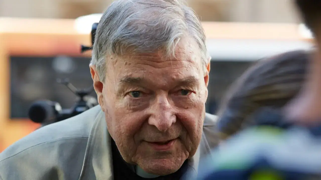 Nahaufnahme von Kardinal Pell, der in die Kamera blickt. Im Hintergrund sind eine Kamera und unscharfe Personen zu sehen.