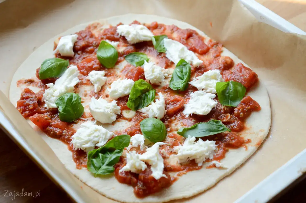 Niedopieczona pizza margherita z sosem pomidorowym, mozzarellą i świeżą bazylią, przygotowana na papierze do pieczenia.