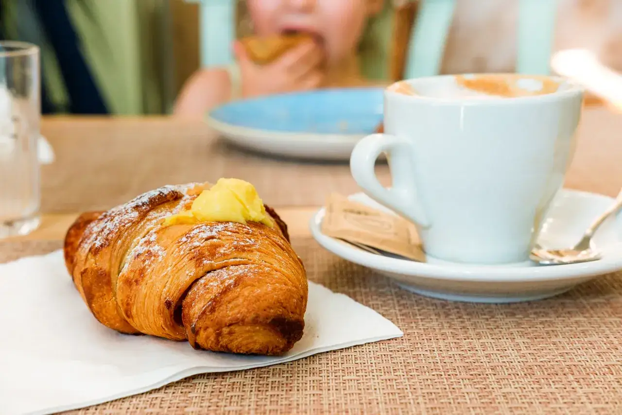 Un delizioso croissant con crema e una tazza di cappuccino, perfetti per una colazione al bar. Sullo sfondo, un bambino mangia qualcosa.