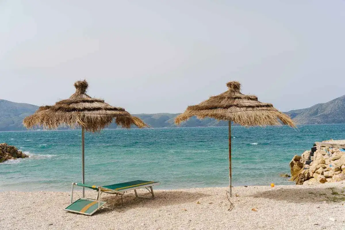 Dwie słomiane parasole i leżaki na kamienistej plaży. Turkusowe morze i góry w tle. Czy Albania to bezpieczny kraj na wakacje? Wygląda spokojnie.