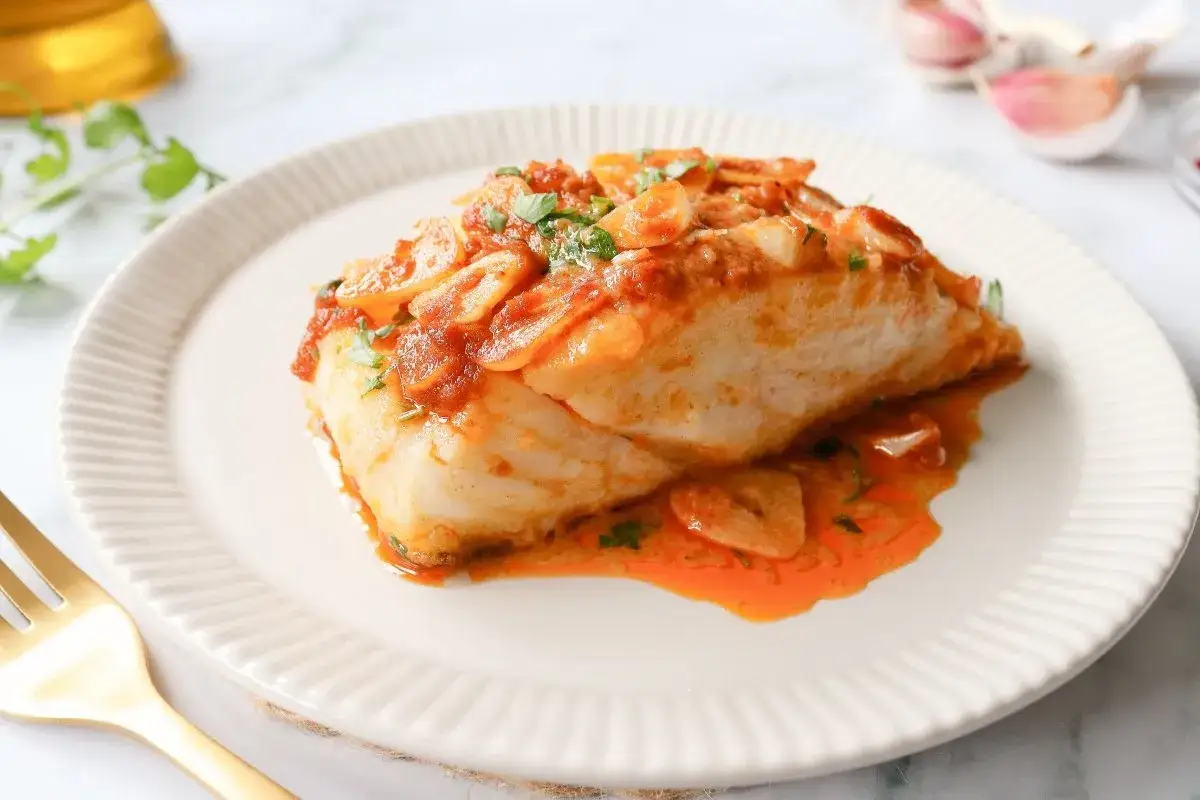 Delicioso bacalao a la llauna con salsa de tomate y ajo, servido en plato blanco con tenedor dorado.