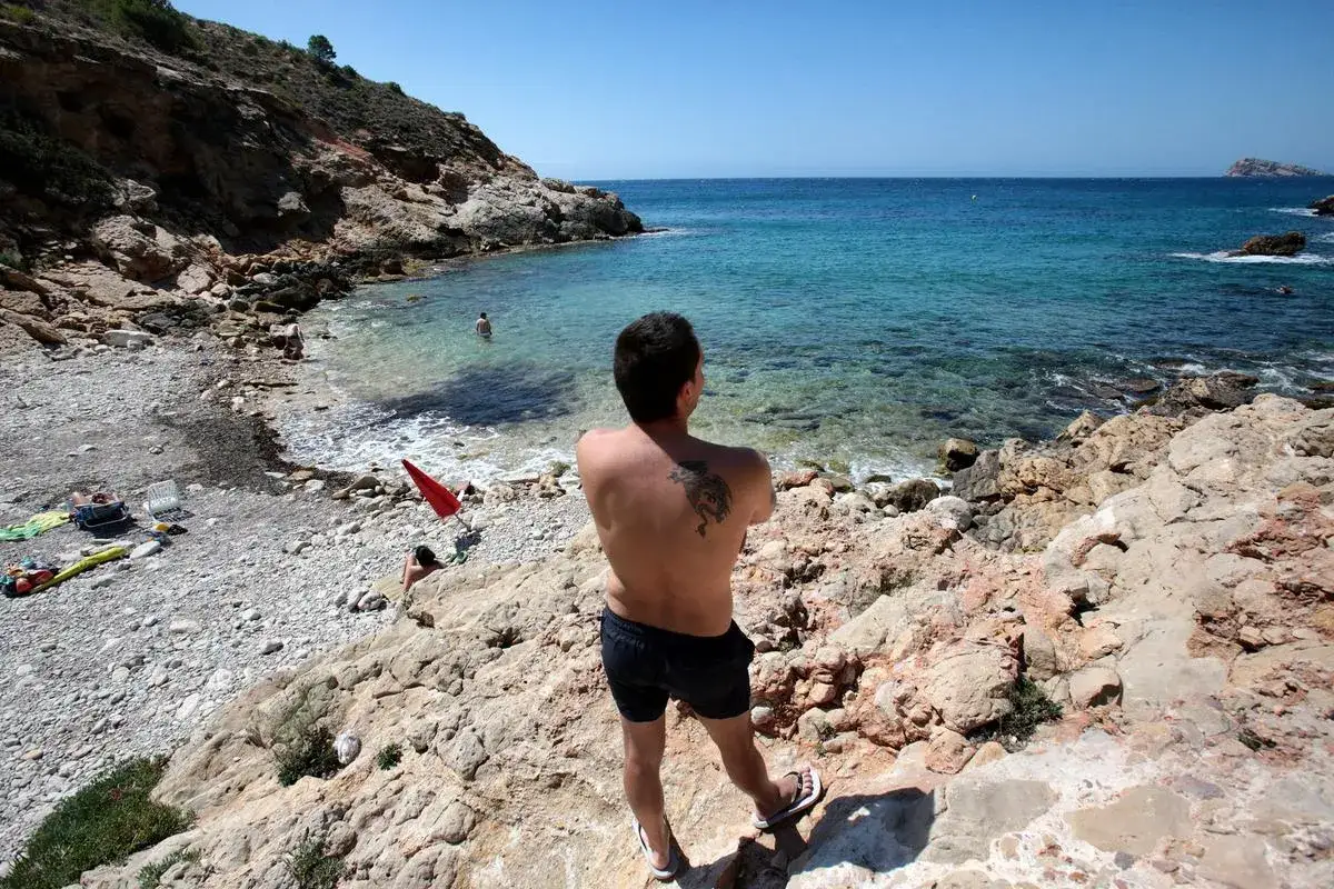 Hombre con tatuaje en la espalda contempla la cala de la playa nudista Benidorm, con aguas cristalinas y rocas.