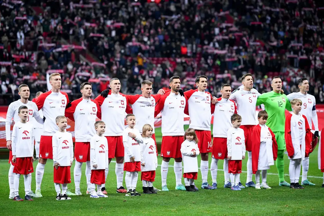 Polscy piłkarze w białych strojach z czerwonymi elementami, obok nich dzieci w strojach klubowych. Stoją na murawie przed trybunami pełnymi kibiców.