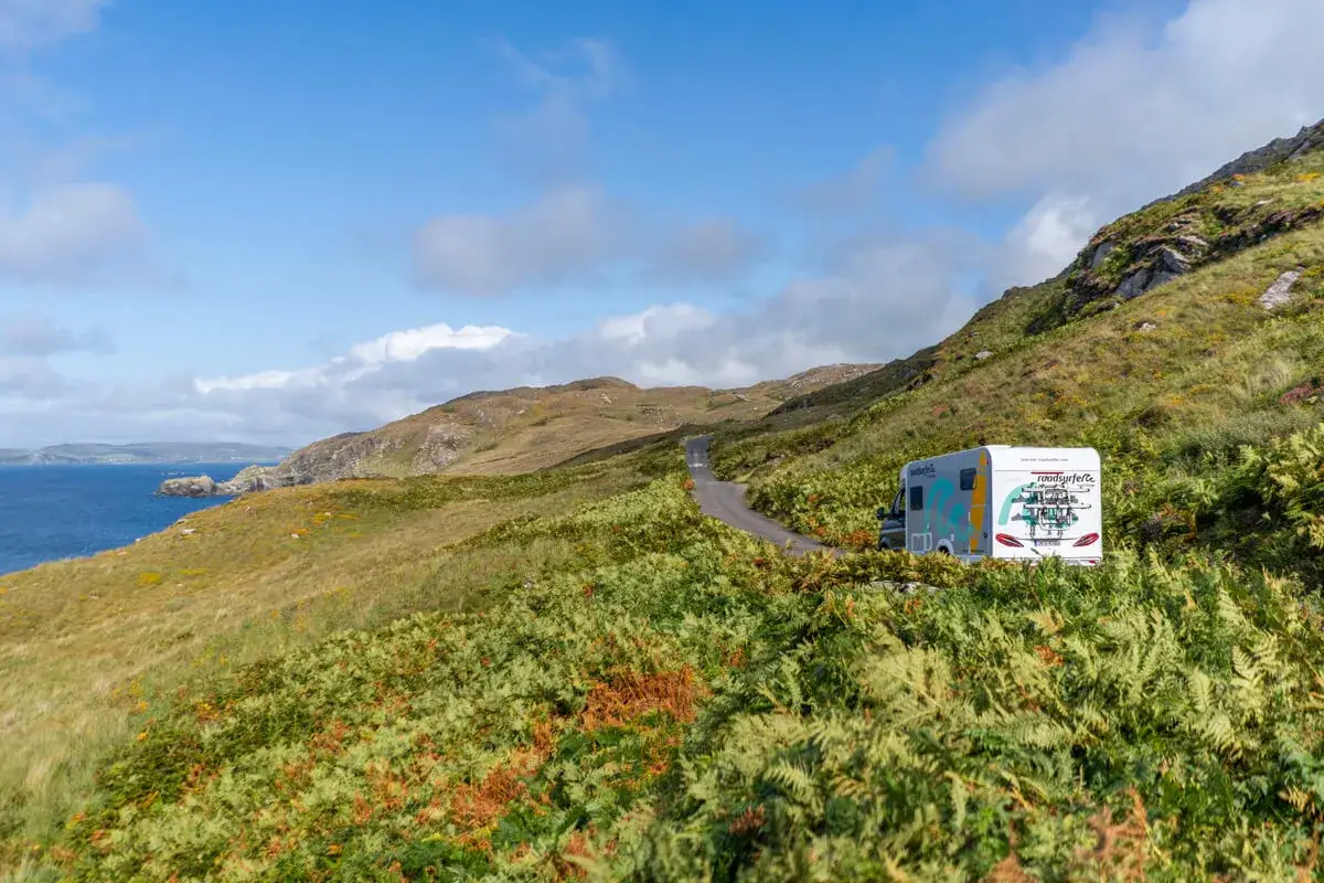 Wohnmobil auf einsamer Straße, umgeben von üppiger irischer Landschaft mit Farnen und dem Meer im Hintergrund.