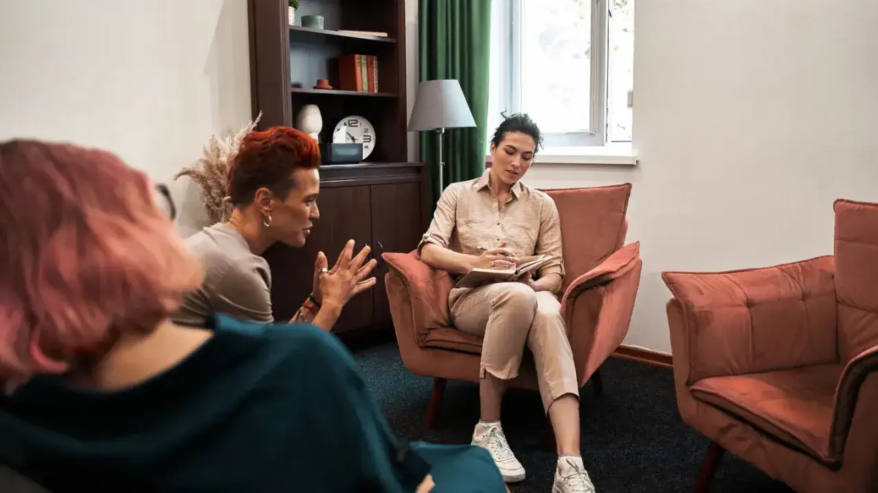 Trzy osoby w gabinecie terapeutycznym. Jedna z nich, z rudymi włosami, gestykuluje, jakby mówiła, że terapia gestalt nie działa.