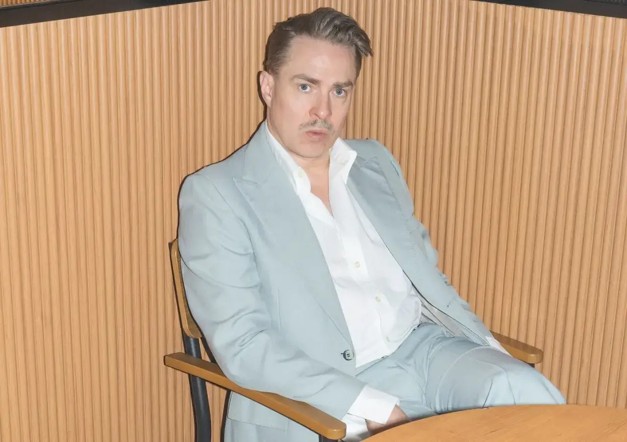Christian Ohlsson sitter i en ljusblå kostym och vit skjorta framför en trävägg.