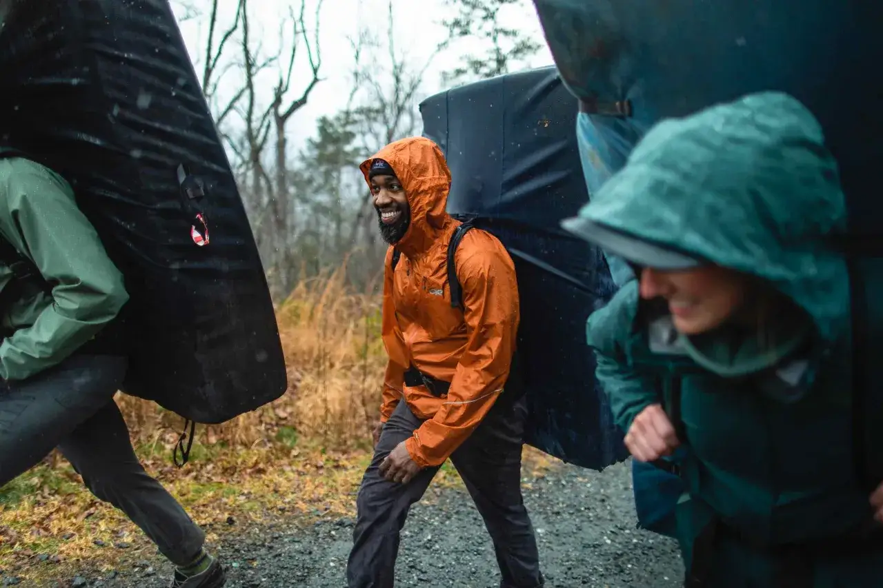 Drei Kletterer tragen dicke Matten durch den Regen. Die orangefarbene Didriksons Jacke des Mannes ist nass, aber er l&auml;chelt.