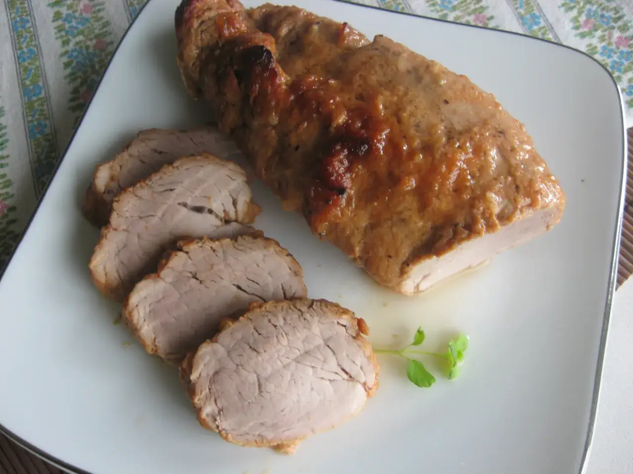 Soczysta polędwiczka wieprzowa pieczona w piekarniku, pokrojona w plastry, podana na białym talerzu z gałązką zieleniny.