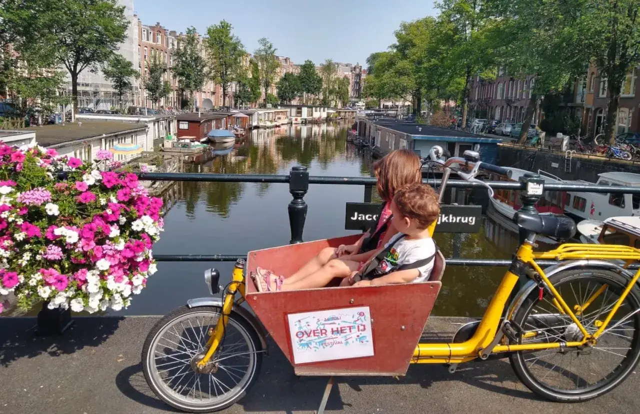 Dos niños disfrutan de un paseo en bicicleta de carga por los canales de Holanda, rodeados de casas flotantes y flores vibrantes.