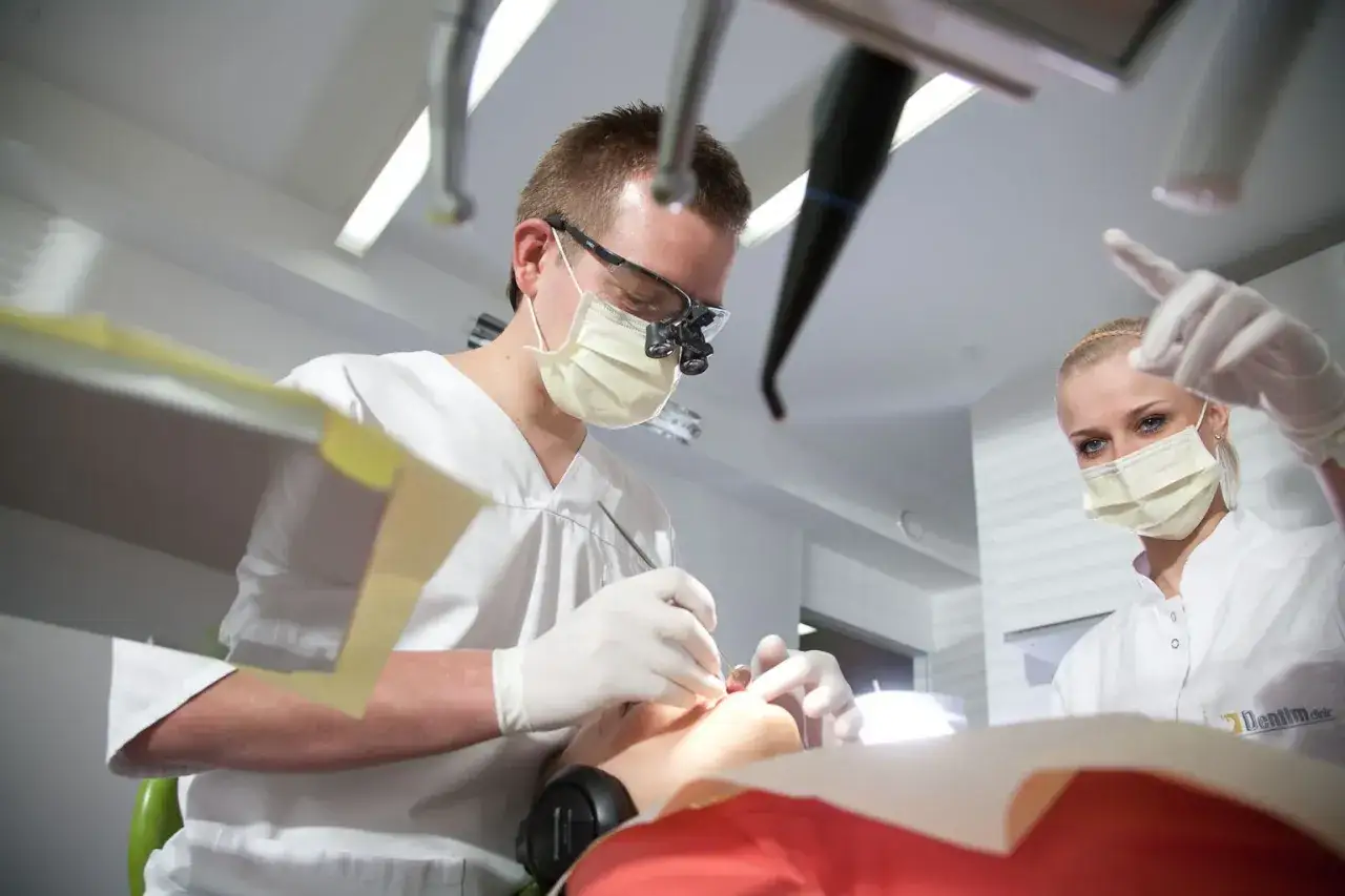 Dentysta w okularach i masce wykonuje zabieg kiretażu. Asystentka w masce wskazuje coś palcem.