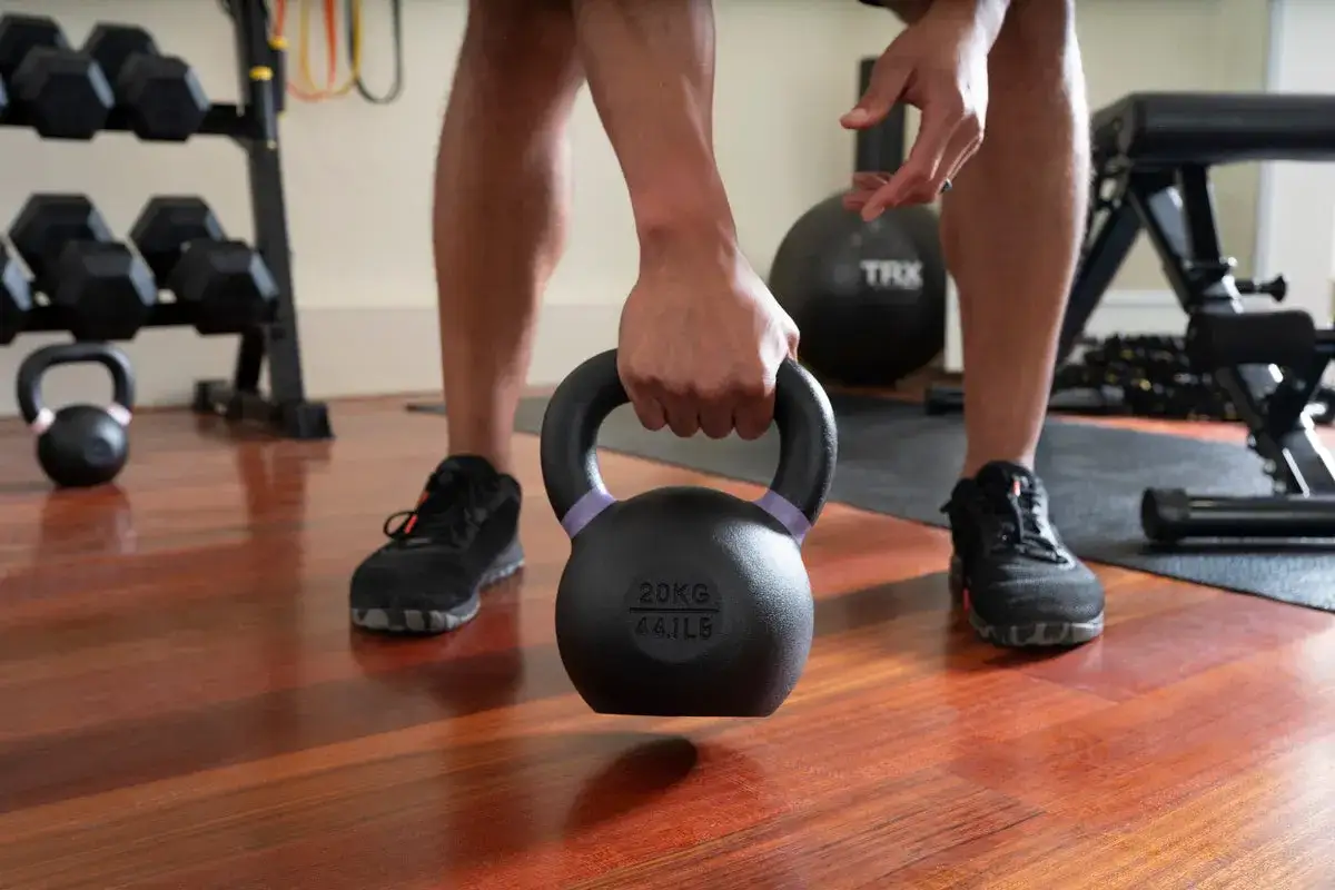 Mann hebt Kettlebell f&uuml;r Brusttraining. Hantelst&auml;nder und Trainingsmatte im Hintergrund.