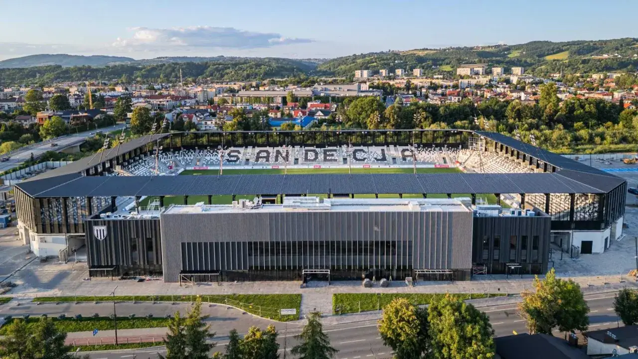 Nowoczesny stadion Sandecji z trybunami i herbem klubu. W tle miasto i wzgórza.