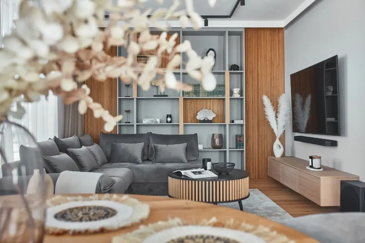 Nowoczesna metamorfoza salonu: szara sofa, okrągły stolik, regał z dekoracjami i drewniane panele.
