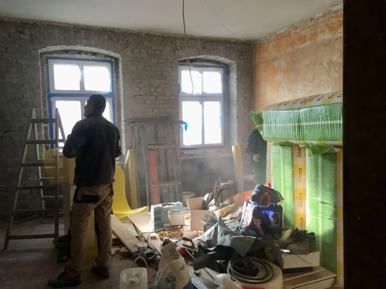 Remont pokoju z dwoma oknami, materiałami budowlanymi i pracownikami.