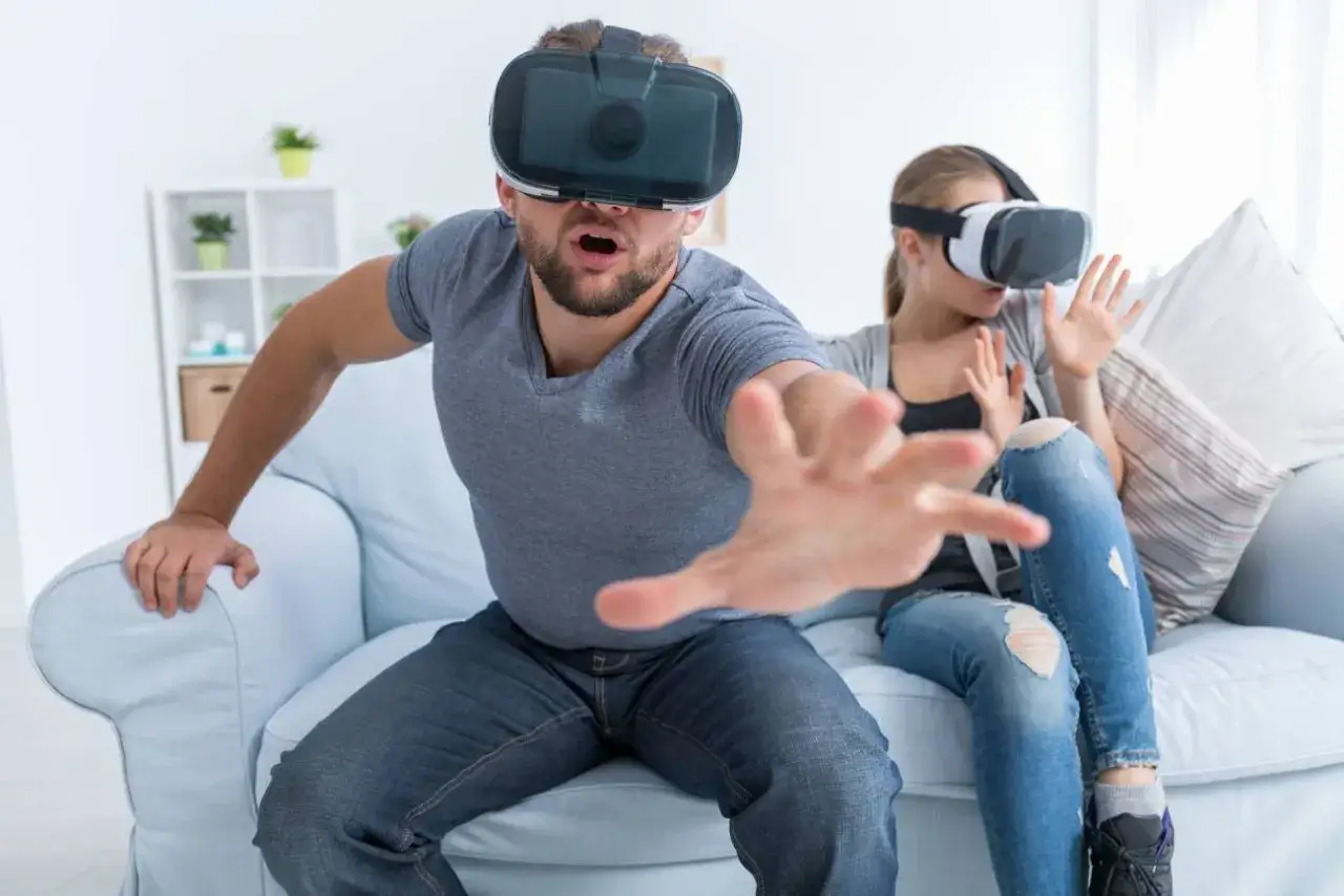 Para w goglach VR przeżywa emocje podczas gry. Mężczyzna wyciąga rękę, jakby łapał coś wirtualnego.