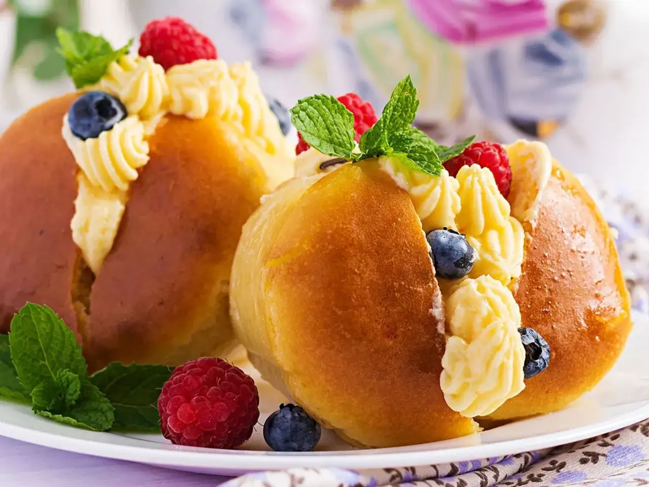 Dos baba postre esponjosos rellenos de crema pastelera y decorados con frambuesas, arándanos y hojas de menta.