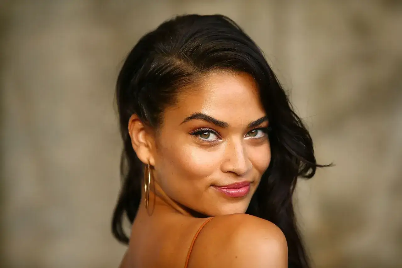 Portrait d'une femme au regard captivant, sa peau de couleur de peau olive rayonne sous la lumière. Ses cheveux sombres encadrent son visage, et elle porte de grandes boucles d'oreilles dorées.