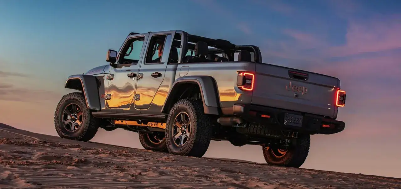 Srebrny jeep pickup z otwartym dachem jedzie po piaszczystej wydmie o zachodzie słońca.