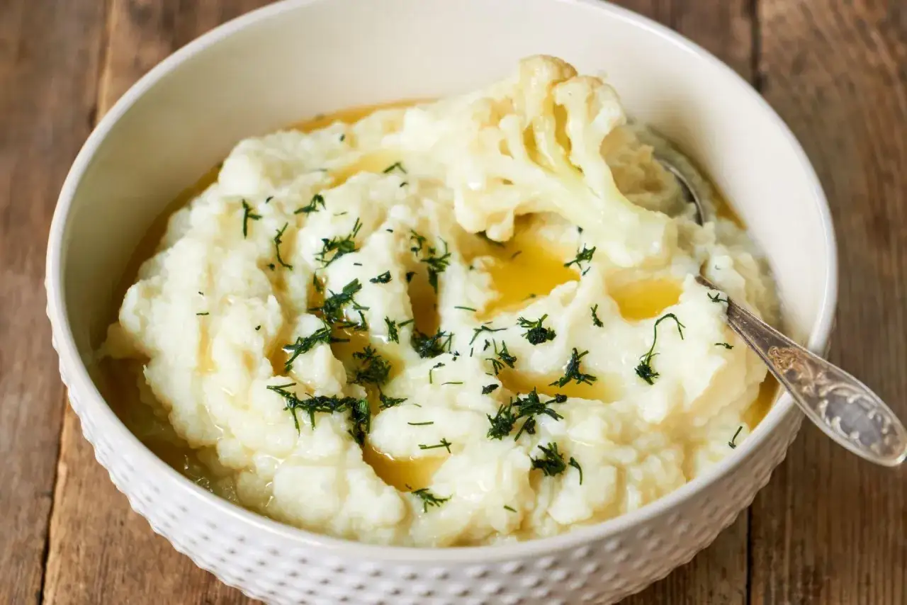 Aksamitne puree z kalafiora, polane masłem i posypane koperkiem, w misce na drewnianym tle.
