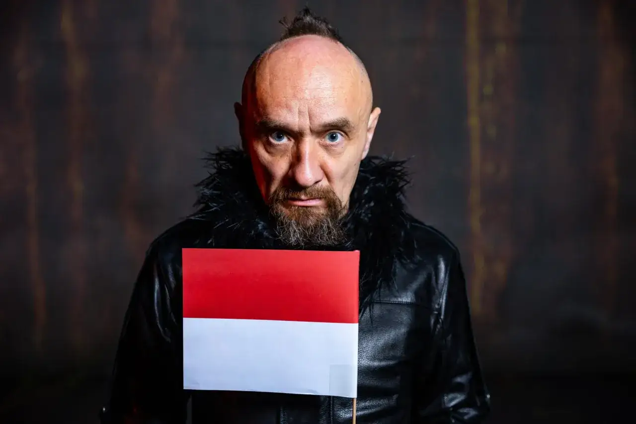 Jan Klata w czarnej sk&oacute;rzanej kurtce z futrzanym kołnierzem trzyma polską flagę. Jego spojrzenie sugeruje emocje towarzyszące spektaklom.