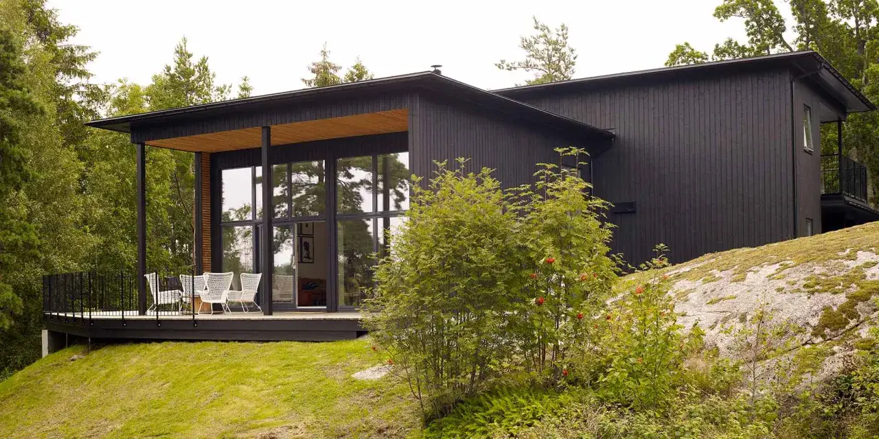 Modernt hus med m&ouml;rkbrun fasad, stora f&ouml;nster och en altan med vita stolar. Omgiven av gr&ouml;nskande tr&auml;d och en mossbelagd sten.