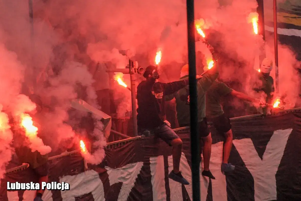 Psycho fans odpalają race, tworząc czerwony dym i chaos. Widać ich sylwetki na tle płomieni.