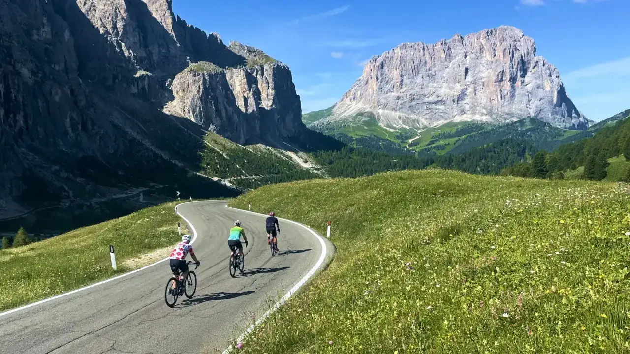 Three cyclists enjoy a scenic cycling holiday in the Dolomites, winding along a mountain road through lush green meadows and past dramatic peaks.