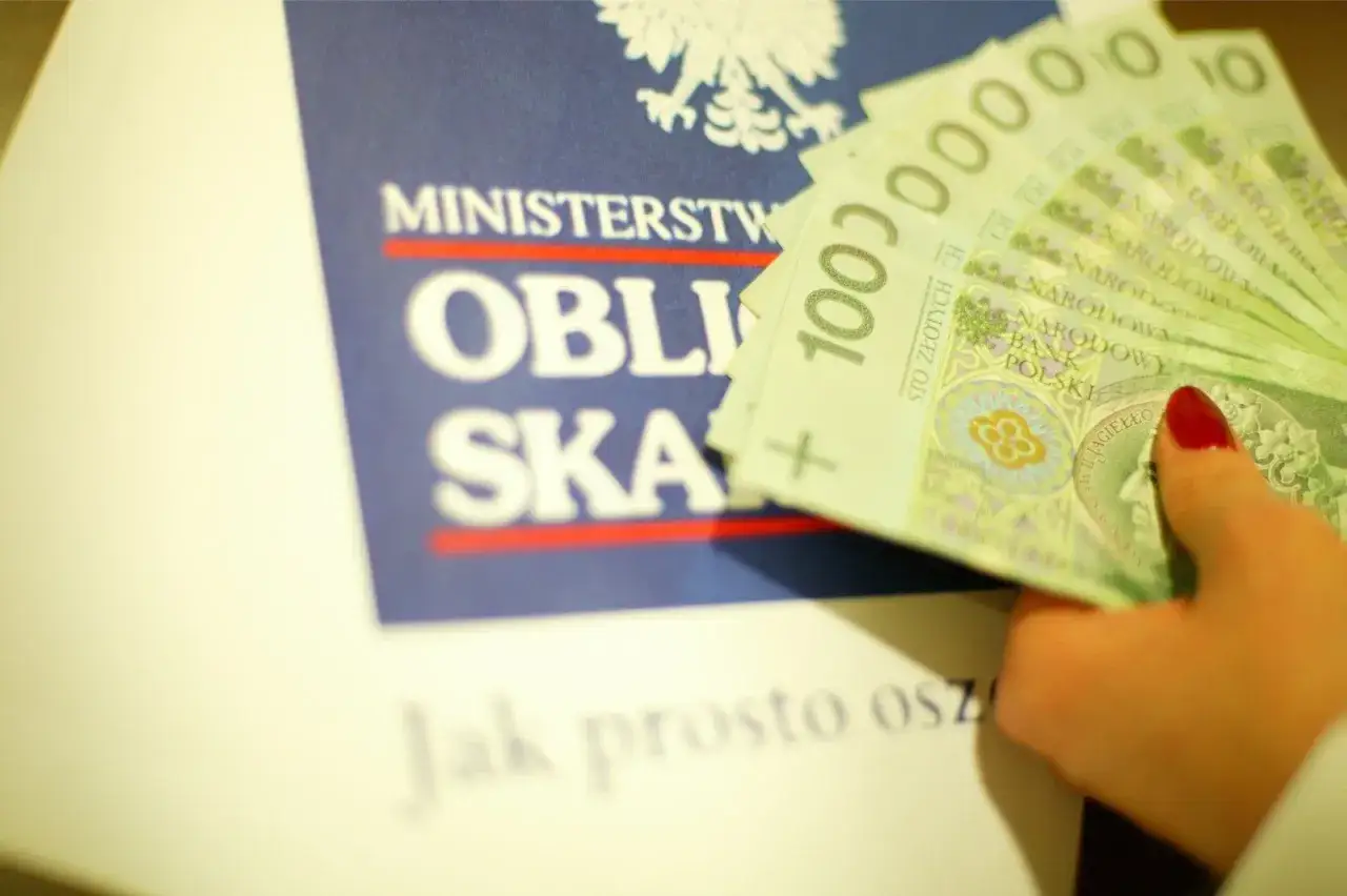 Dłoń z czerwonym lakierem do paznokci trzyma stos banknotów 100 zł na tle napisu