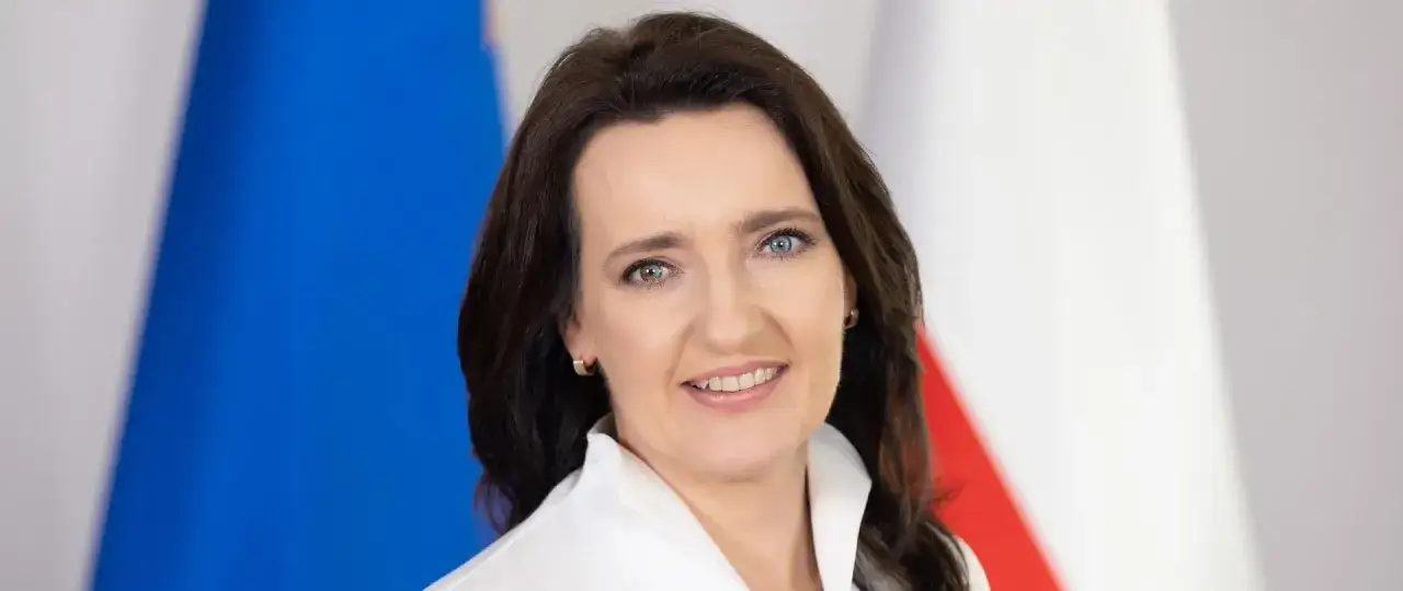 Marzena Okła-Drałowicz, uśmiechnięta kobieta z ciemnymi włosami, na tle biało-czerwonej flagi.