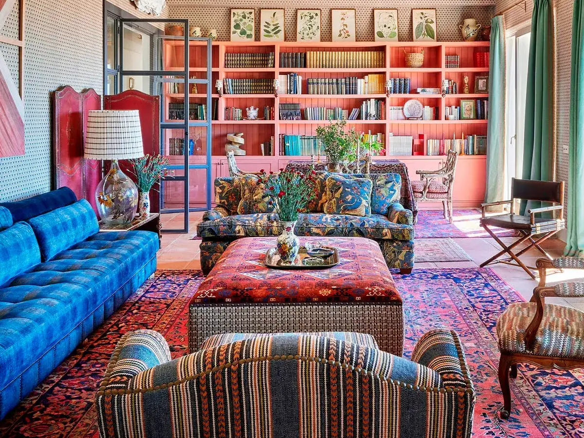 Salón con sofás de terciopelo azul y estampado floral, alfombras persas y estanterías vintage color coral repletas de libros y objetos decorativos.