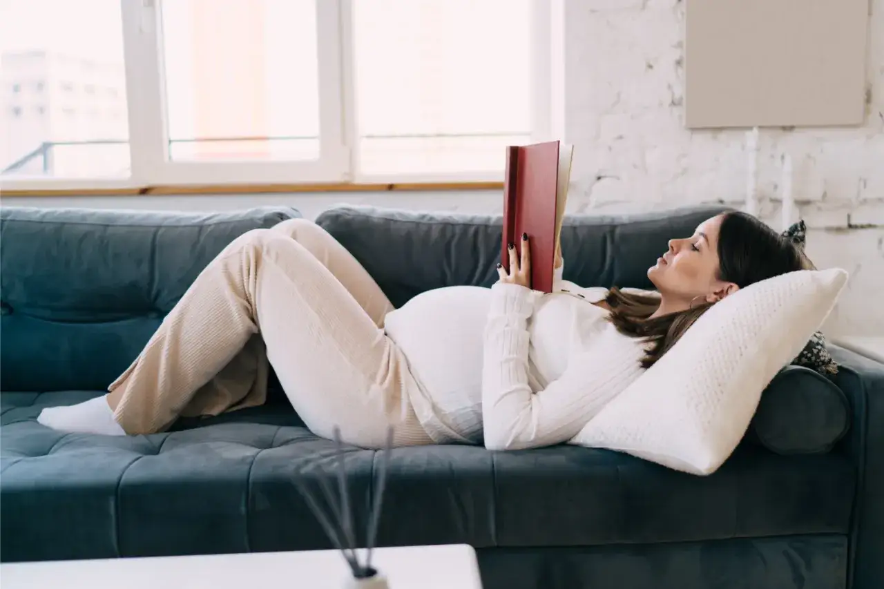 Donna incinta rilassata sul divano legge uno dei tanti libri da leggere in gravidanza, preparandosi all'arrivo del bambino.