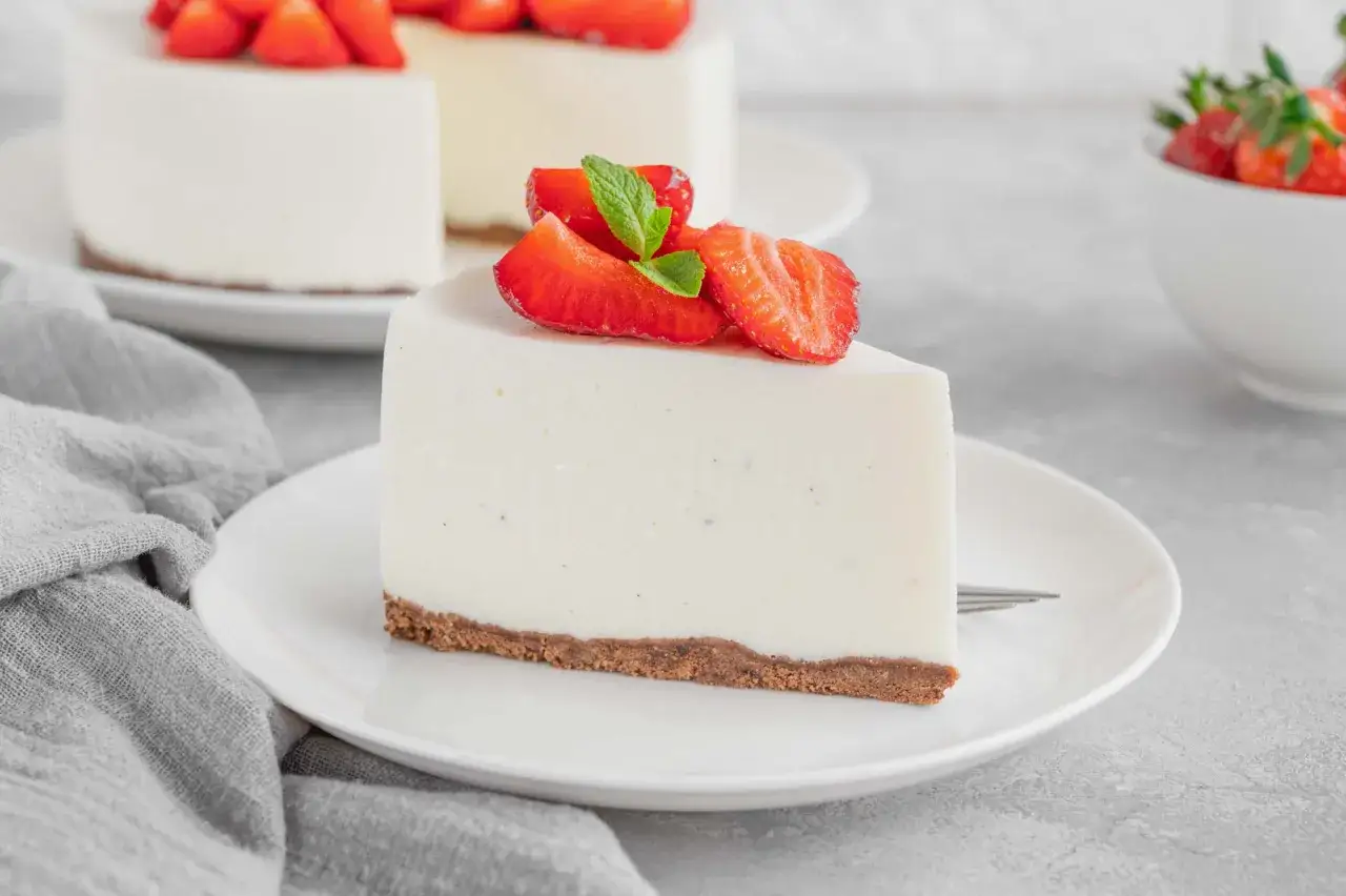 Una fetta di cheesecake con yogurt greco, guarnita con fragole fresche e menta, su un piatto bianco.