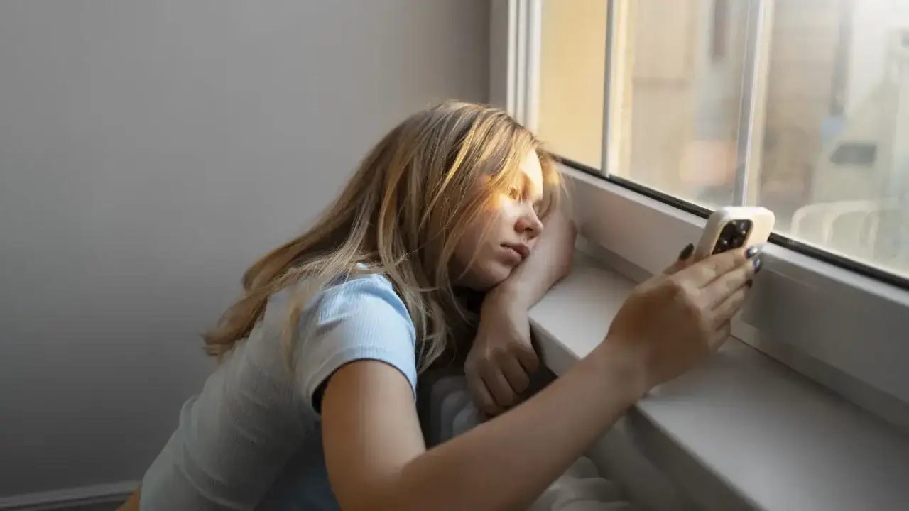 Chica rubia con m&oacute;vil, mirando por la ventana, pensativa. Quiz&aacute;s buscando frases cuando no te responden.