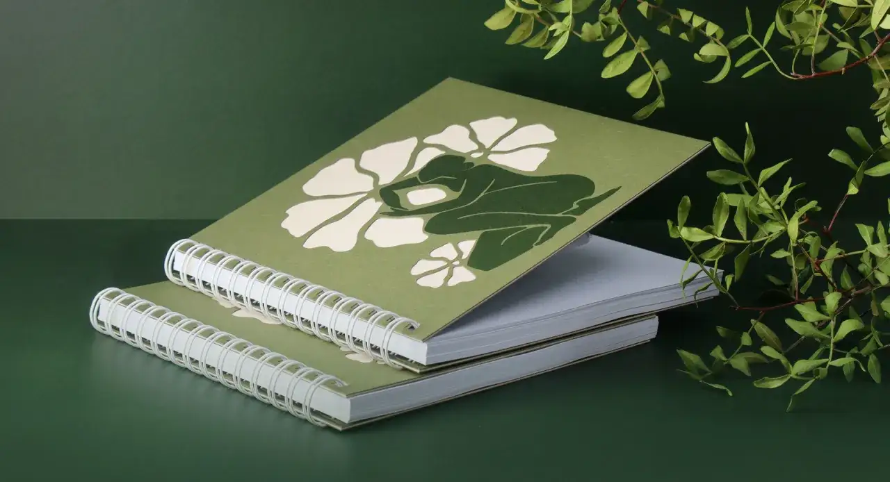 Dos cuadernos en espiral con una figura verde y flores blancas. La impresión gráfica evoca la naturaleza.