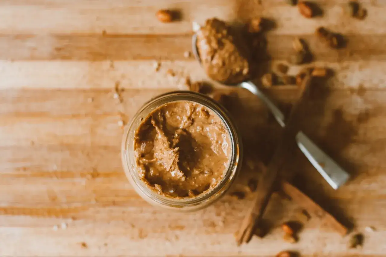 Glas mit cremiger Erdnussbutter, Löffel und Zimtstangen auf Holzbrett. Erdnussbutter gesund und lecker.