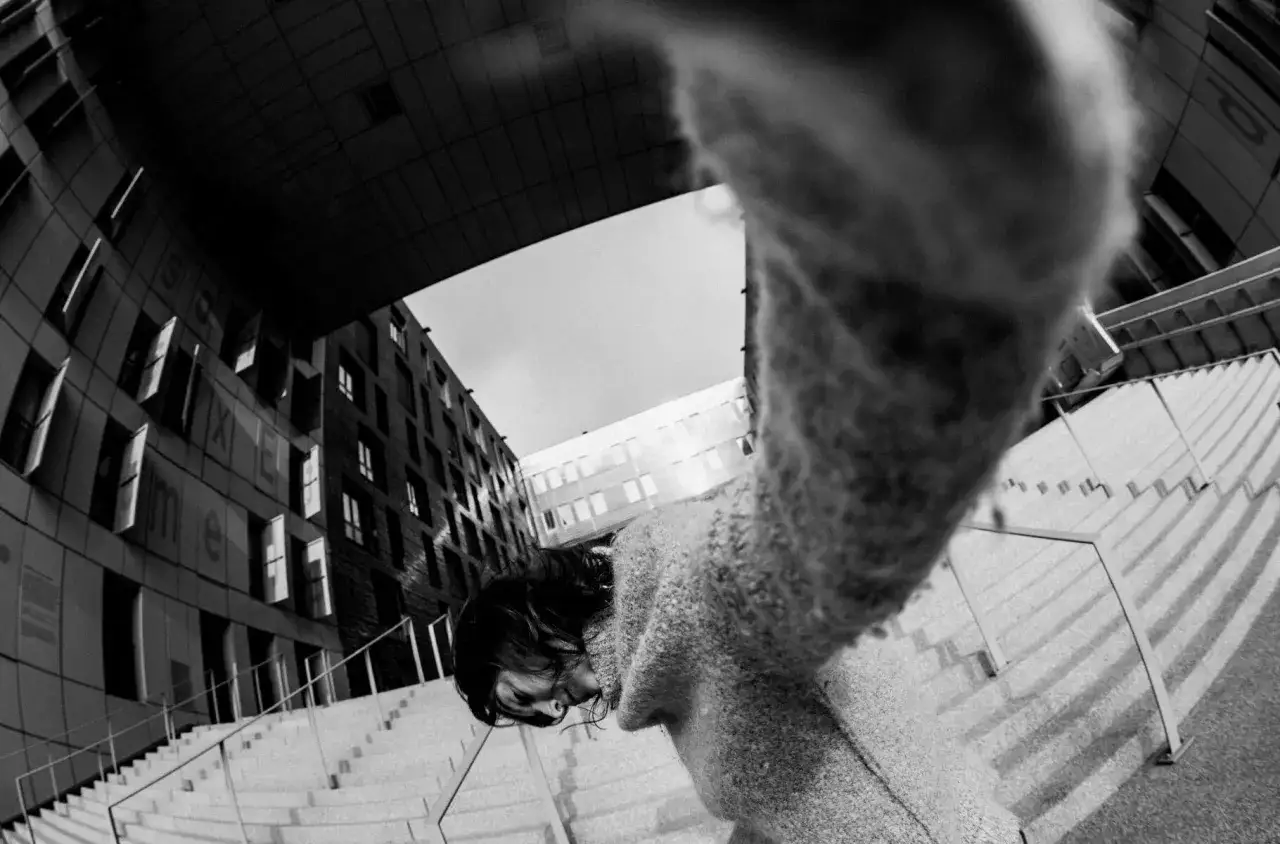 Osoba w swetrze pochyla się nad schodami, a szerokokątny obiektyw tworzy efekt rybie oczy, zniekształcając architekturę.
