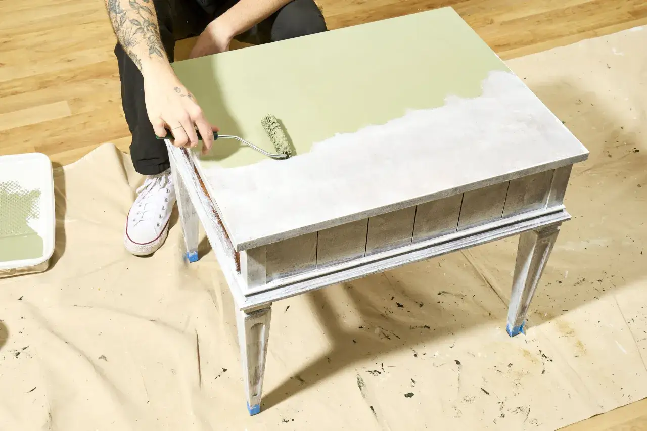 Persona con tatuajes pinta con rodillo una mesa de madera de color verde pálido, renovando su aspecto.
