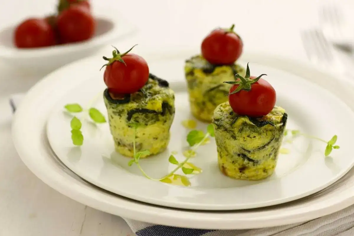 Tre deliziosi flan di zucchine, ognuno sormontato da un pomodorino fresco, guarniti con foglioline verdi e un filo d'olio.