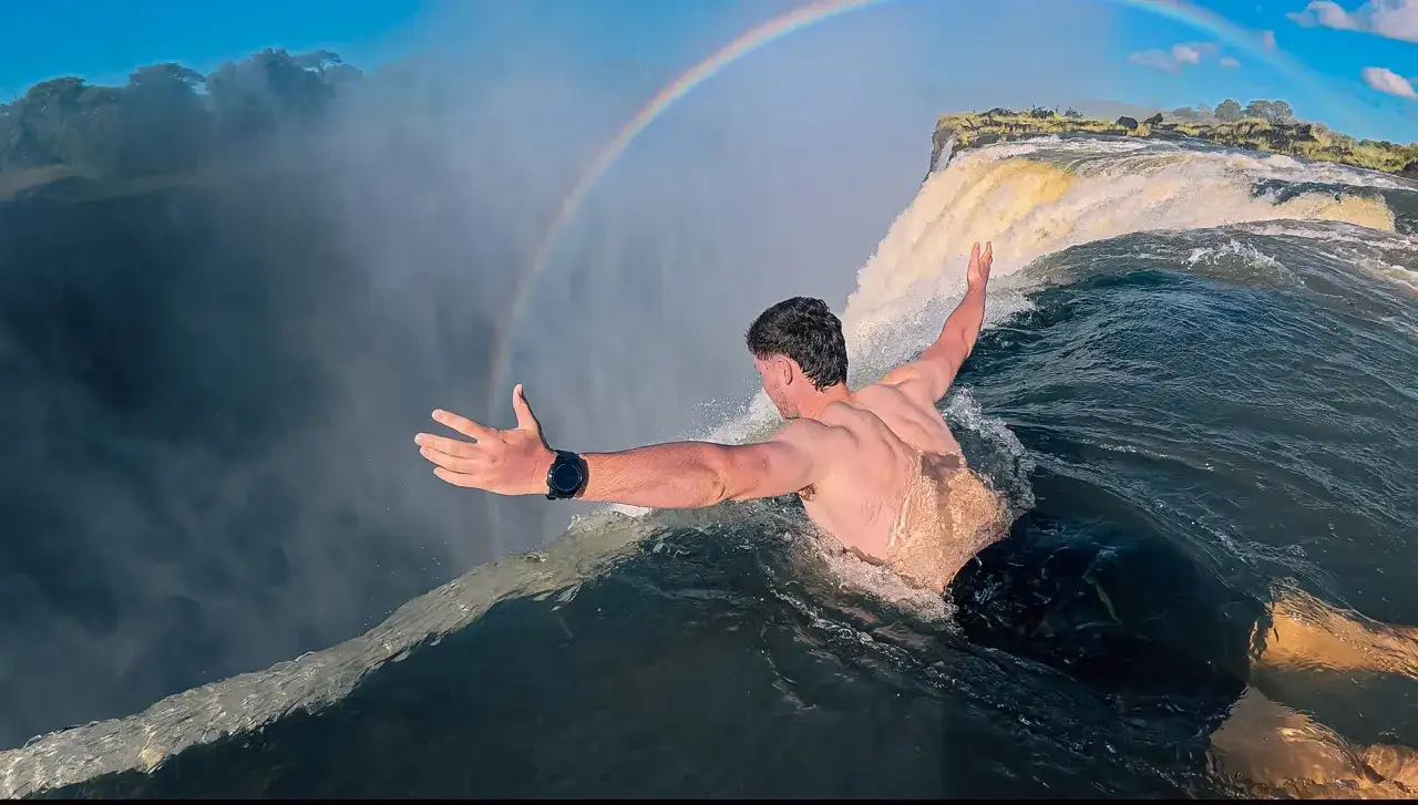 Mann schwimmt am Rand des Devils Pool, mit einem Regenbogen über den Victoriafällen.