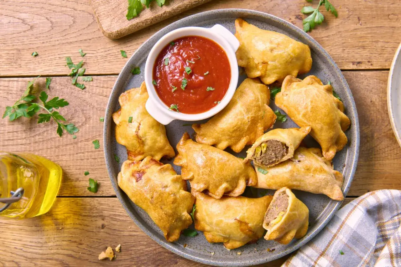 Złociste empanadas z mięsnym farszem, podane z sosem pomidorowym i świeżą natką pietruszki na drewnianym stole.