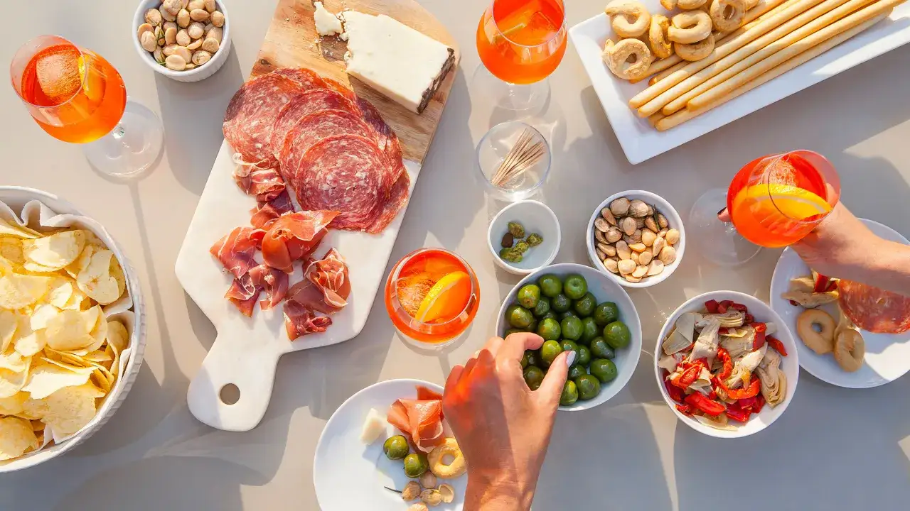 Włoski aperitif: kieliszki z pomarańczowym drinkiem, deska z wędlinami i serem, miseczki z oliwkami, orzechami, chipsami i suszonymi pomidorami.