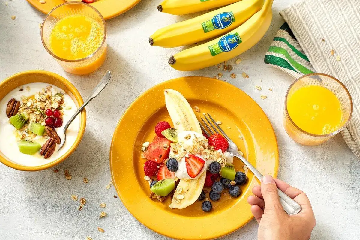 Una banana a colazione, tagliata a met&agrave; e guarnita con yogurt, frutta fresca e granola. Accanto, una ciotola di yogurt e un bicchiere di succo d'arancia.