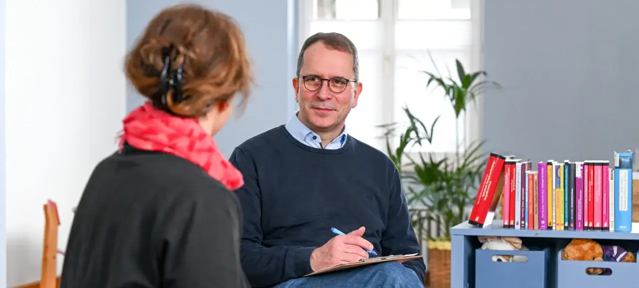 Ein Mann mit Brille und einem blauen Pullover spricht mit einer Frau. Er h&auml;lt einen Stift und Notizen. Auf dem Regal stehen viele B&uuml;cher, die zeigen, dass liebe ist eine entscheidung.