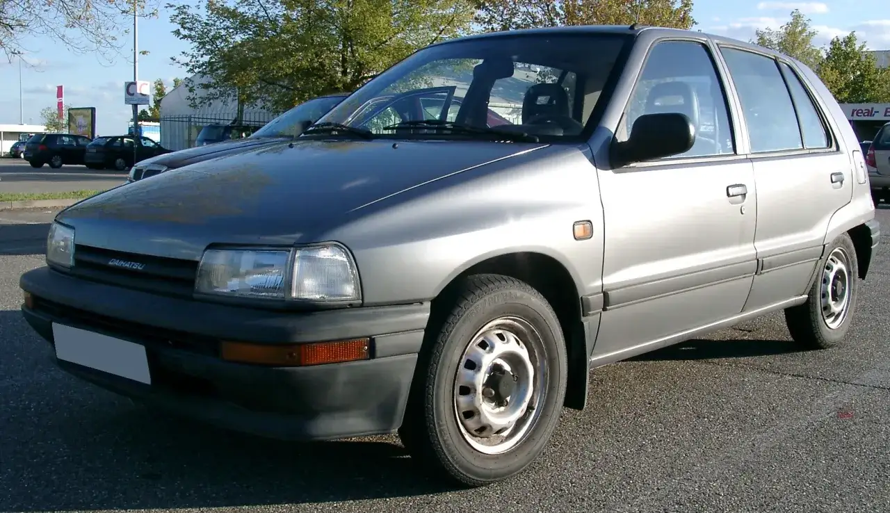 Srebrny Daihatsu Charade na parkingu, z widocznymi drzewami i budynkami w tle.