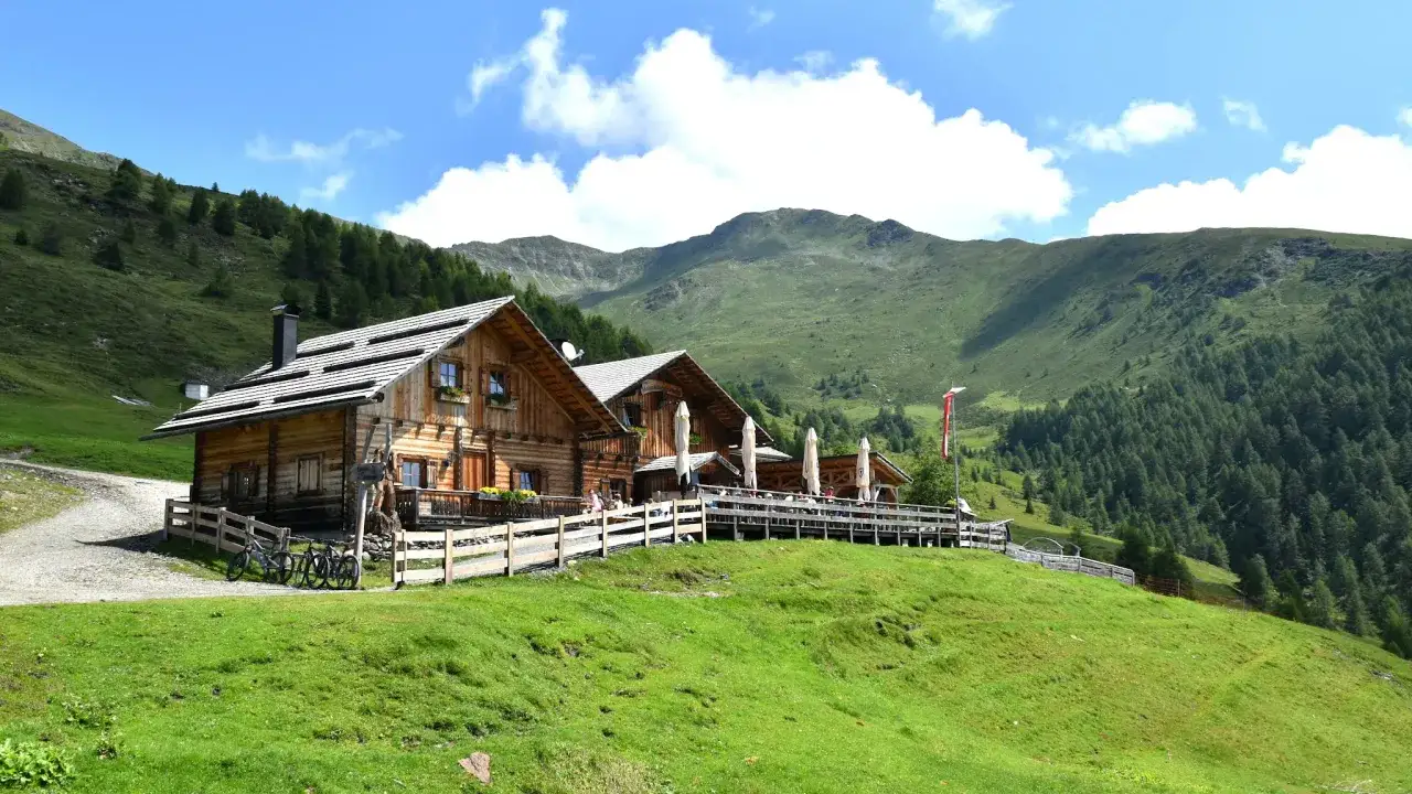 Rifugio alpino in legno con terrazza panoramica e tavoli all'aperto, circondato da prati verdi e boschi, con montagne sullo sfondo. I forni di sopra sono spenti, ma l'atmosfera è accogliente.